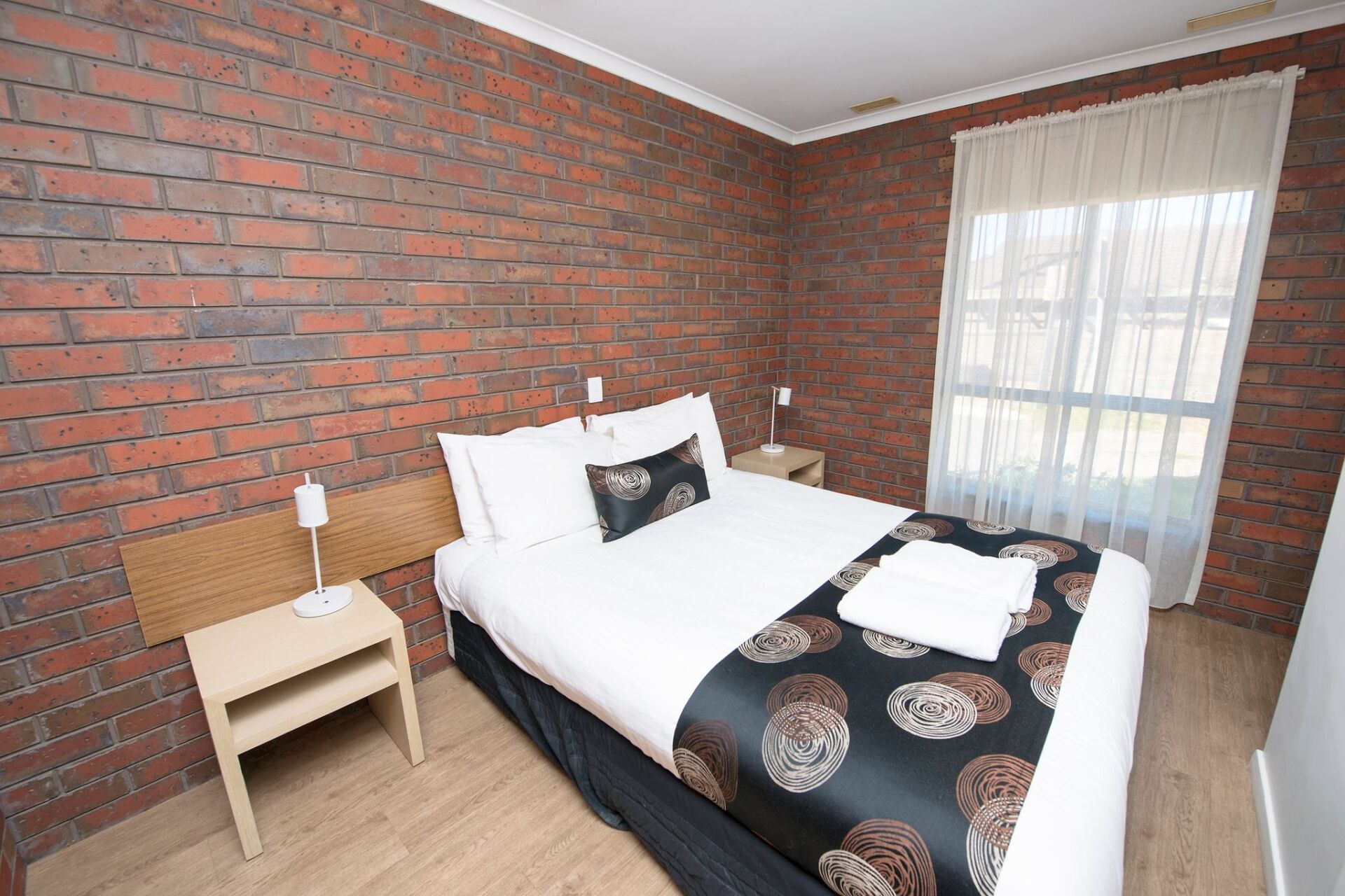 Bedroom with brick wall, double bed, small bedside tables, window with sheer curtain.