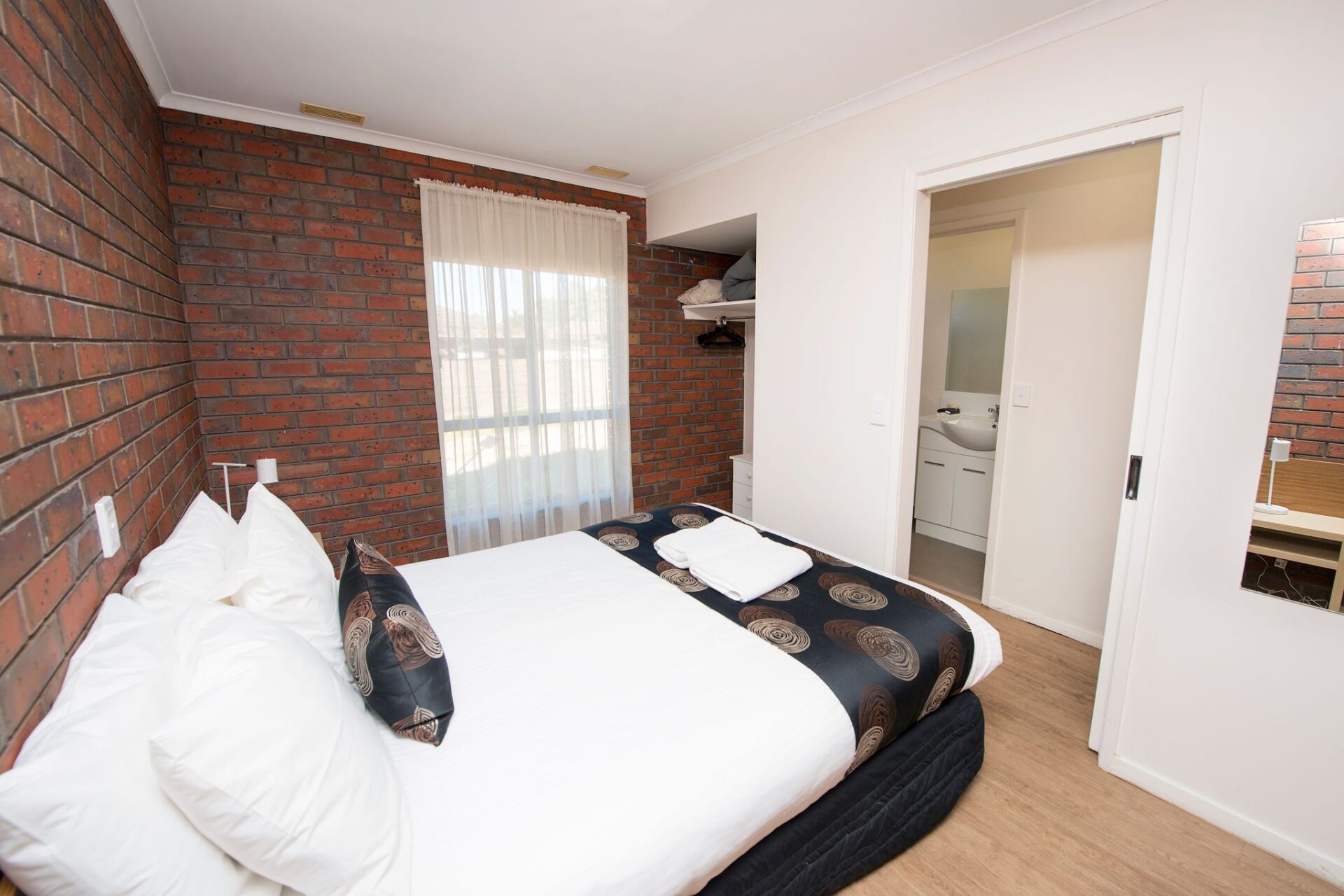 Bedroom with brick wall, double bed, window with curtains, and bathroom door.