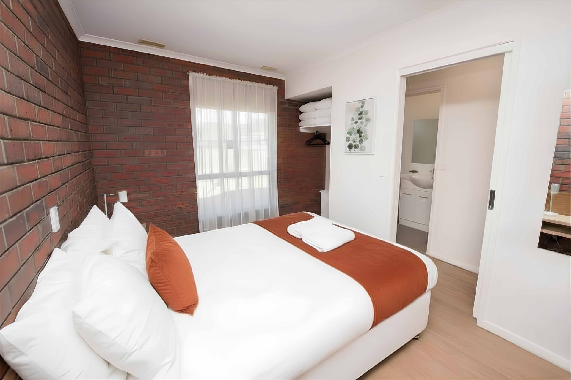Bedroom with brick wall, double bed, window with curtains, and bathroom door.