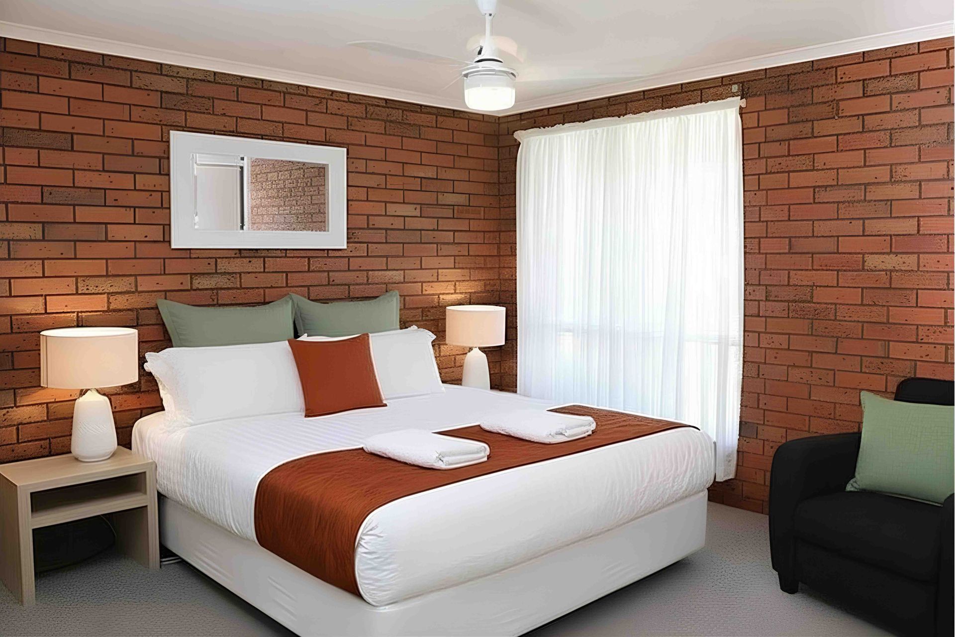 Bedroom with brick wall, double bed, small bedside tables, window with sheer curtain.