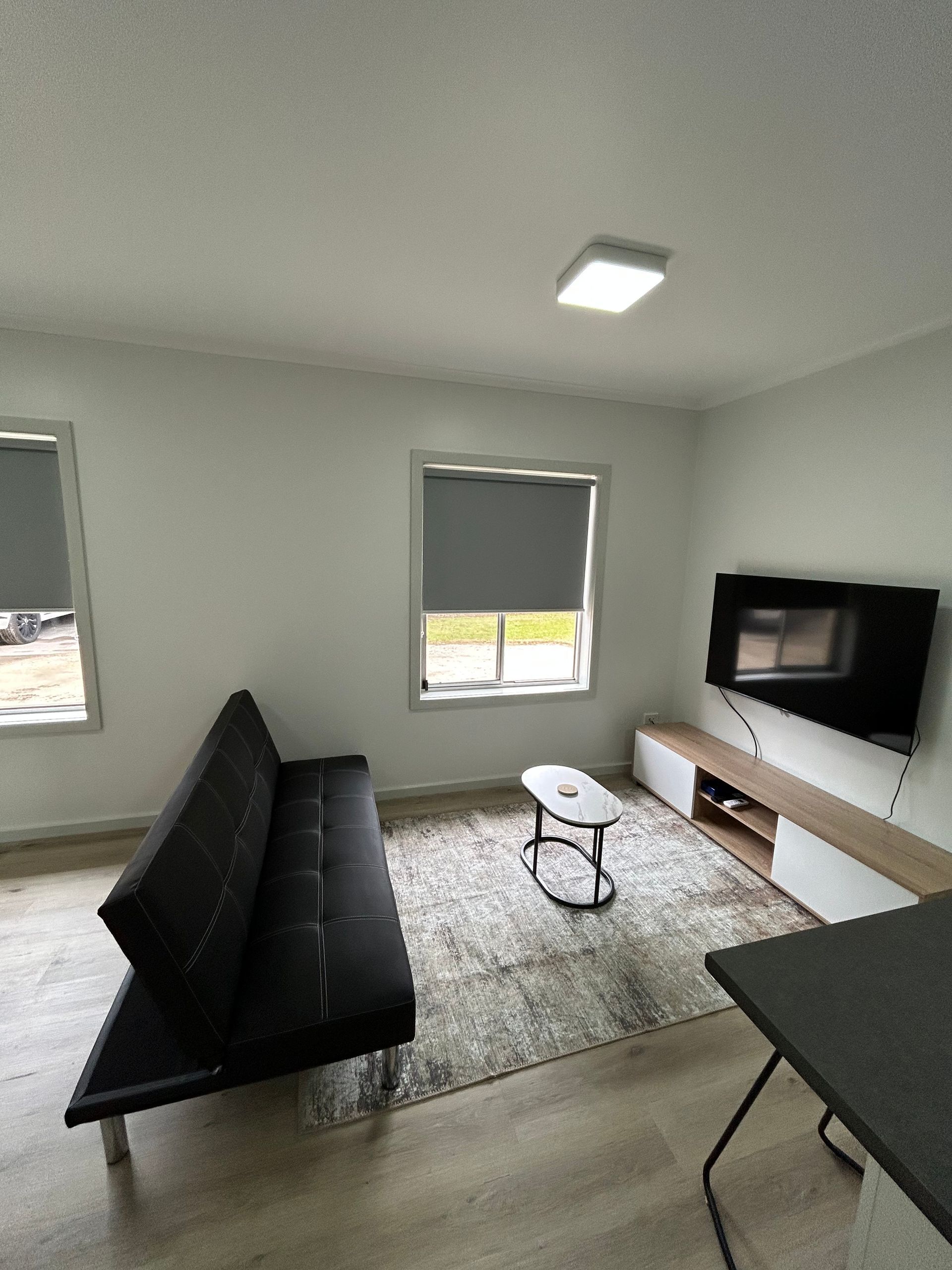 Living room with a black futon, TV, coffee table, and windows with gray shades. Light wood floors and white walls.