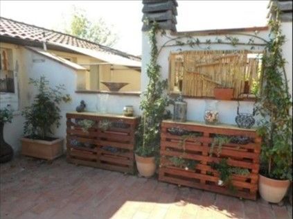 Due fioriere in pallet di legno con piante in vaso sono appoggiate a un muro bianco esterno su un patio in mattoni.