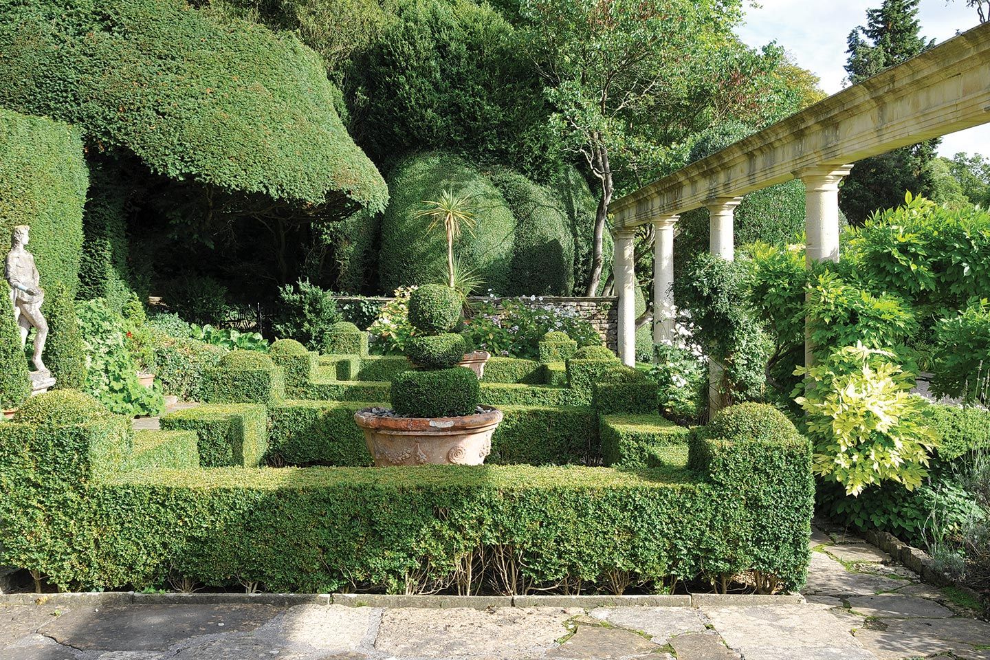 Un giardino formale con siepi di bosso ben curate, una scultura vegetale a più livelli in un vaso di terracotta, un pergolato in pietra e una statua.