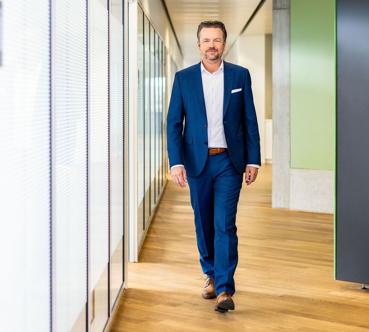 Eine Person in einem blauen Anzug und weißem Hemd geht einen modernen Büroflur mit Holzboden und Glaswänden entlang.