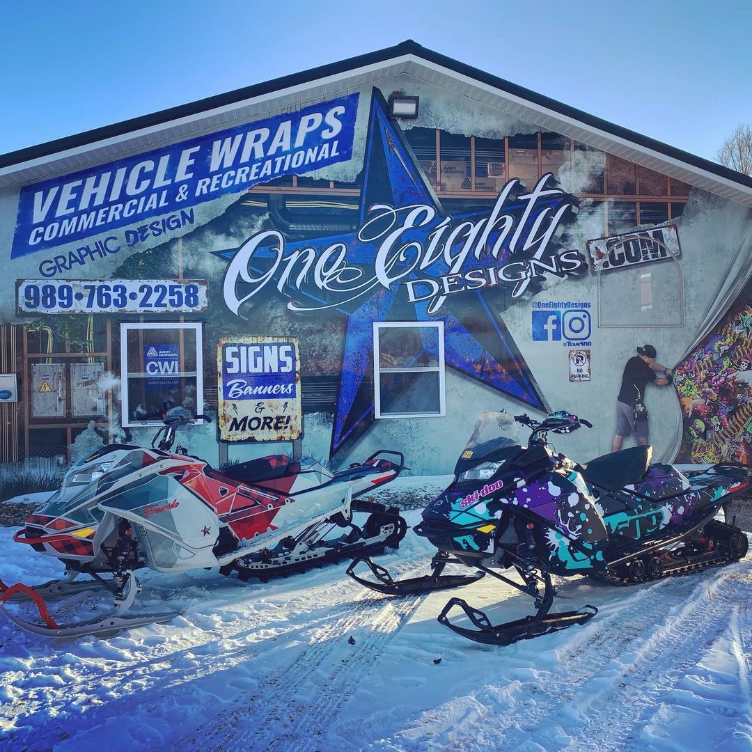 Two snowmobiles are parked in front of a building that says vehicle wraps.