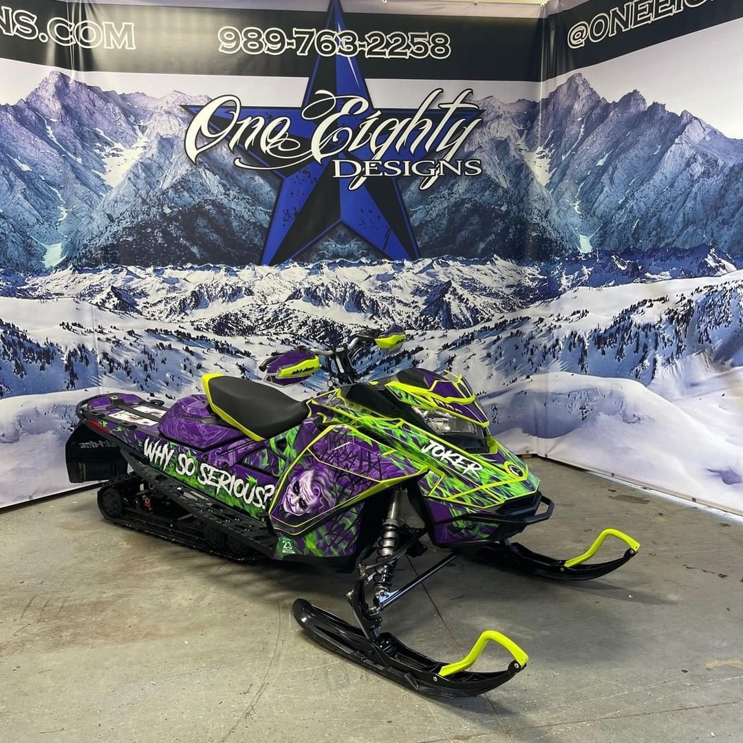 A green and purple snowmobile is parked in front of a sign that says one eighty designs