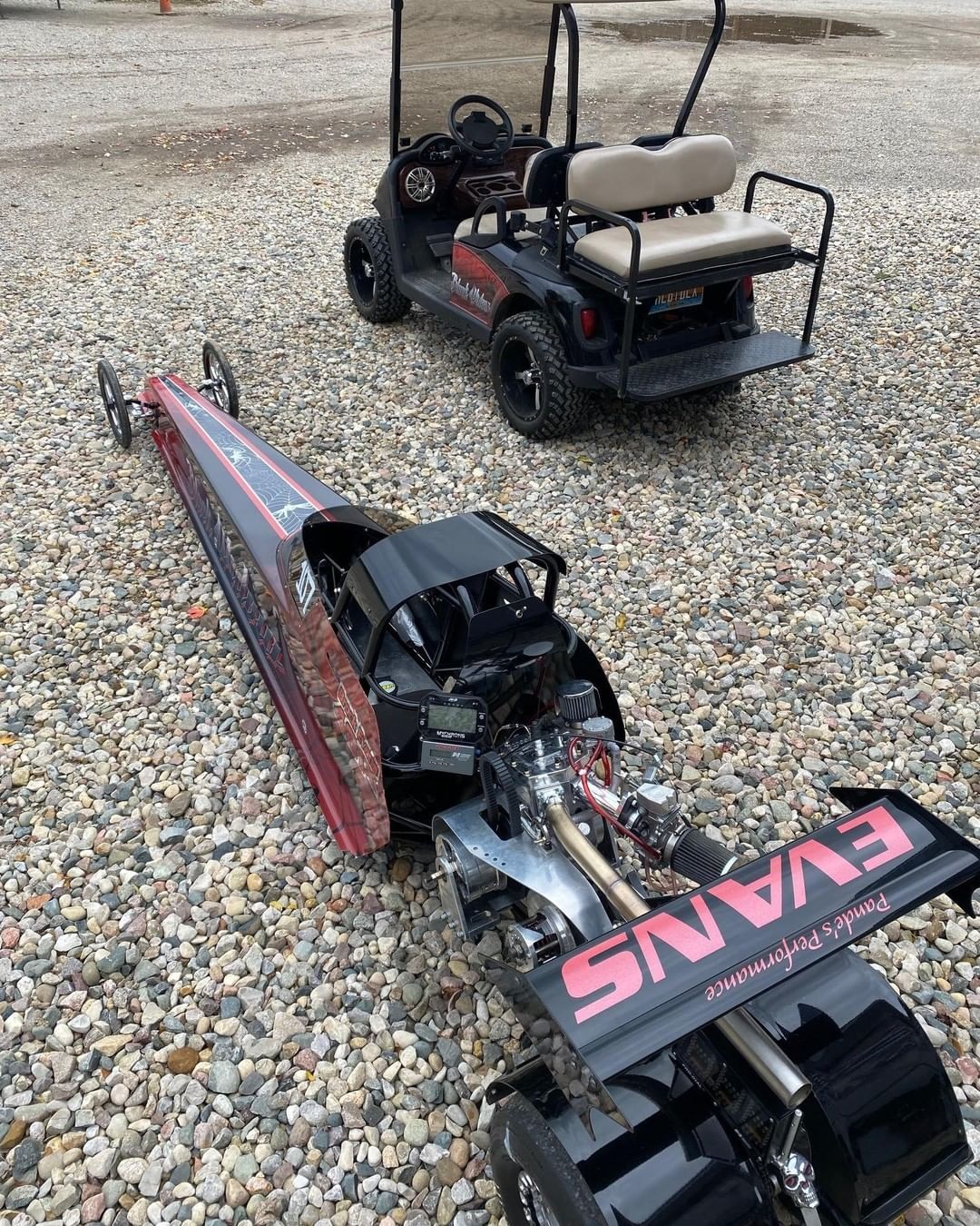 A dragster is sitting next to a golf cart on a gravel road.