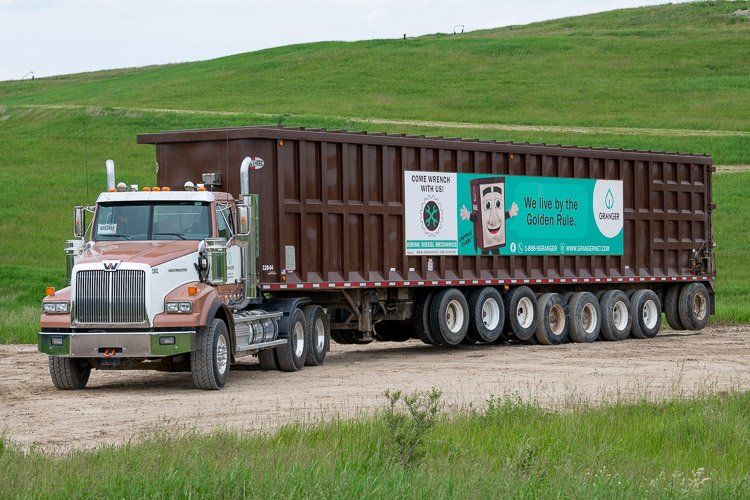 A semi truck is driving down a dirt road next to a grassy hill.