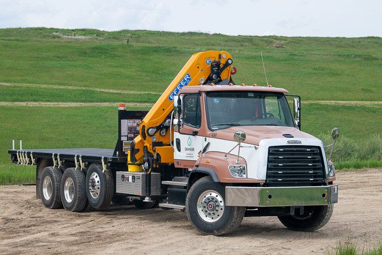 A flatbed truck with a crane on the back is driving down a dirt road.