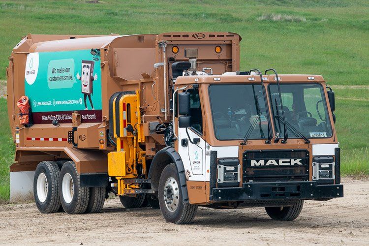 A mack garbage truck is driving down a dirt road