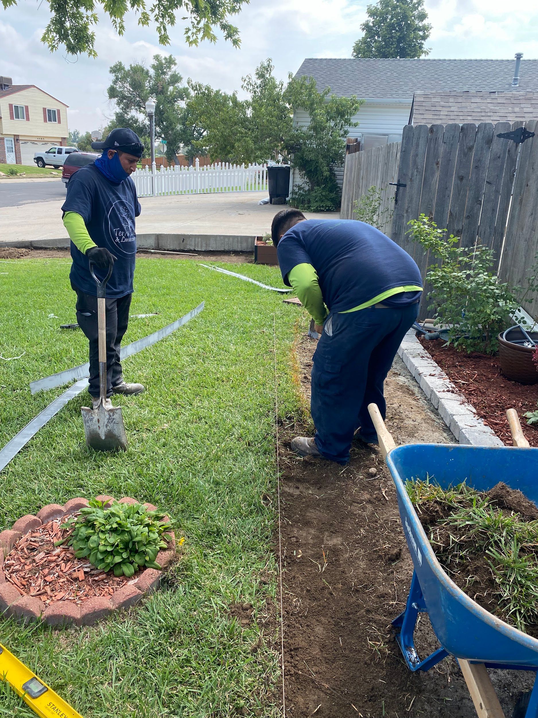 Two workers installing landscaping edging in a front yard. One digs with a shovel, the other uses a level.