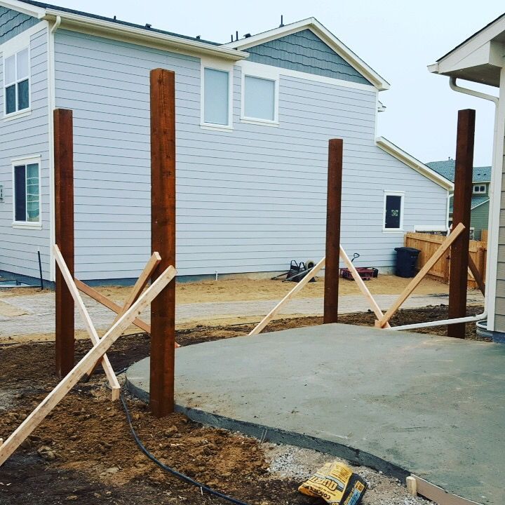 Wooden posts in a concrete patio, braced with wood. A light blue house is in the background under an overcast sky.