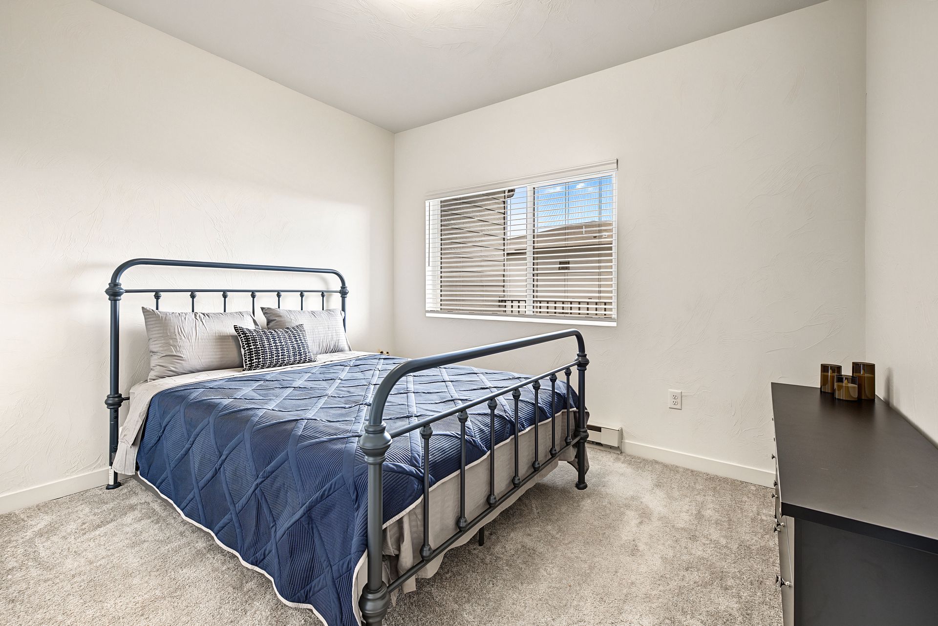 Simple bedroom with a metal bed, blue bedding, dresser, and a window with blinds.
