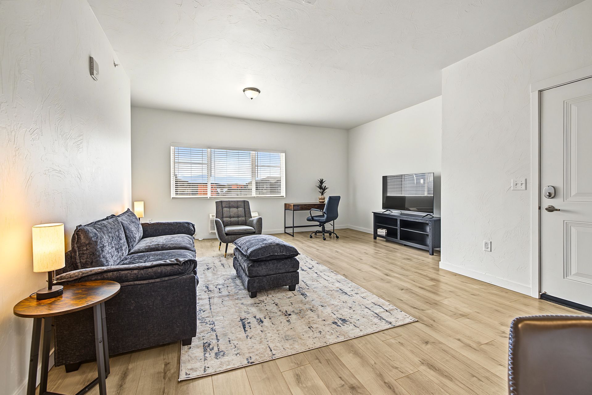 Bright living room with gray sofa, ottoman, TV, desk, and large window on light wood floor.