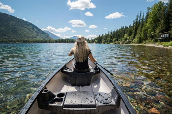 Person paddling a canoe on a clear mountain lake with forested shores and blue sky