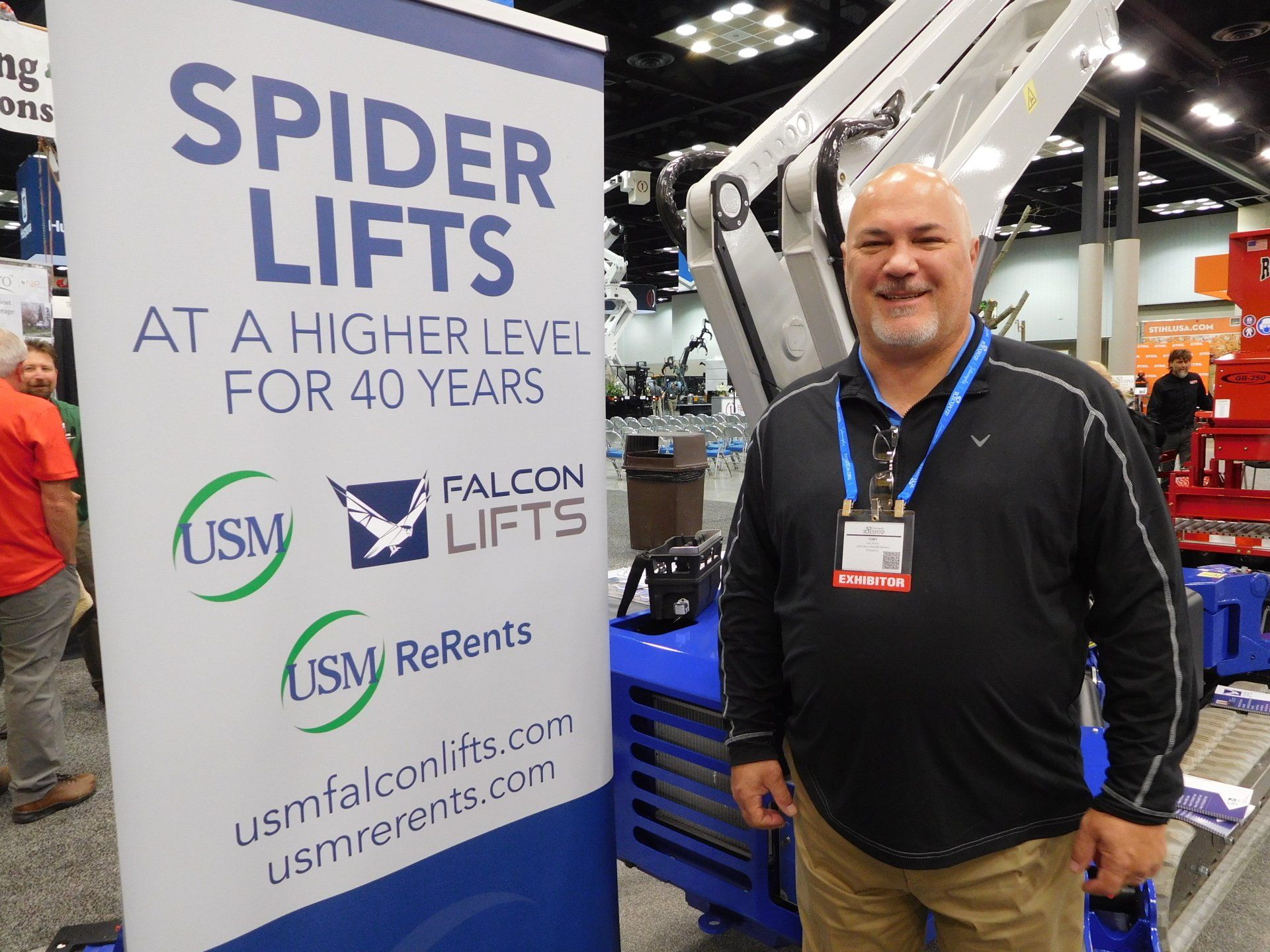 A man is standing in front of a sign that says spider lifts at a higher level for 40 years