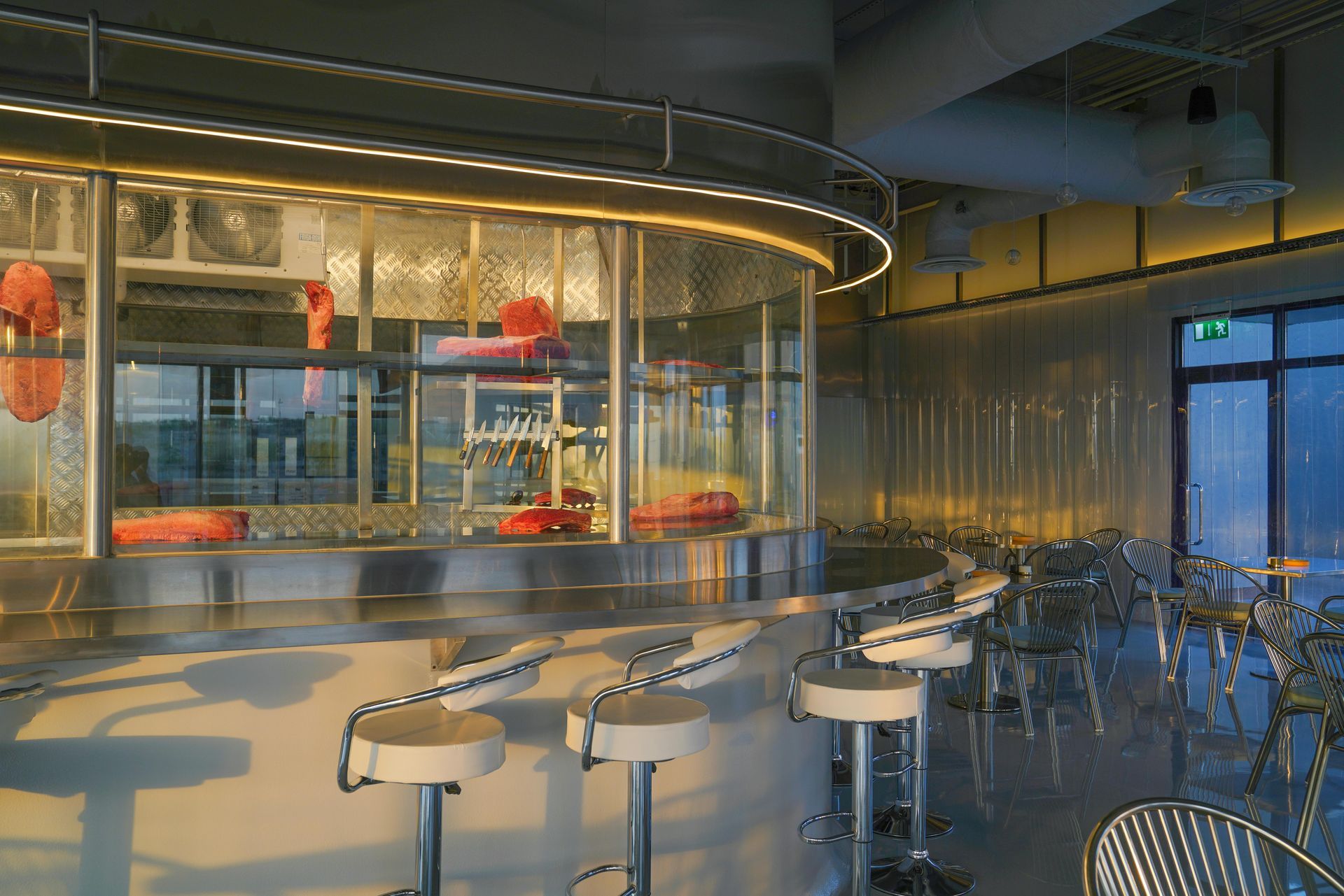A modern, curved stainless steel bar with white swivel stools in a bright, contemporary restaurant interior.