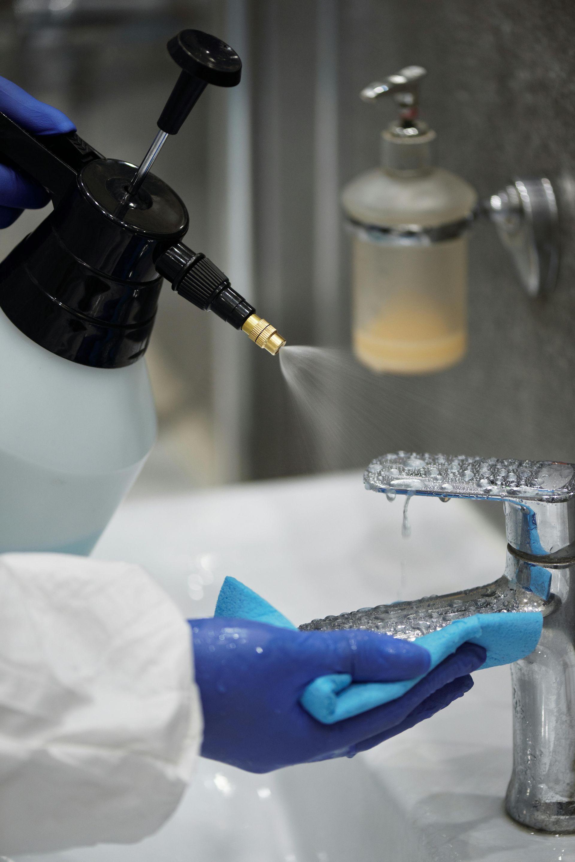 A person in protective gear and blue gloves sprays and wipes a bathroom faucet with a cleaning cloth.