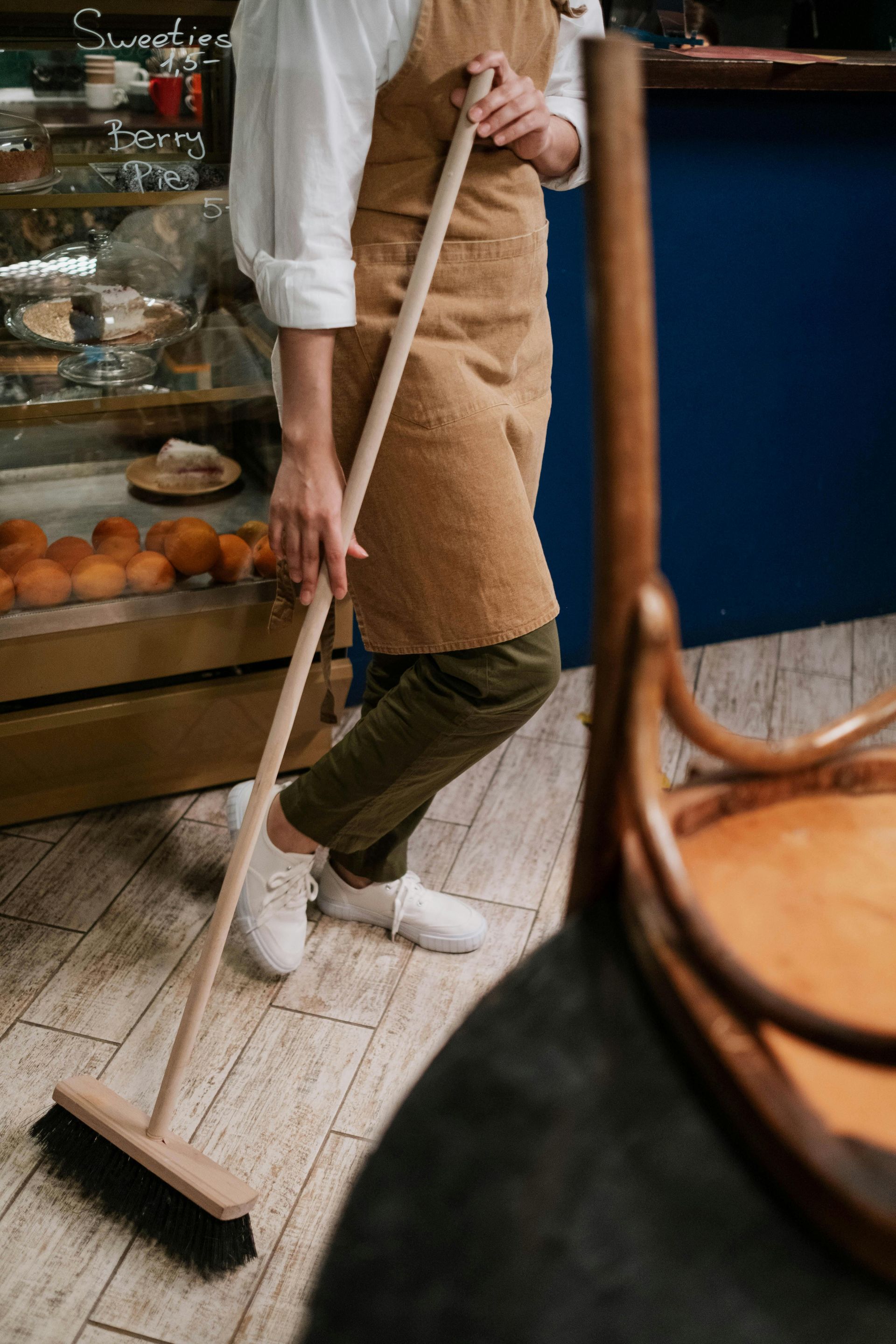 A person in a tan apron and olive pants sweeps a light-colored wood floor in a bakery setting.