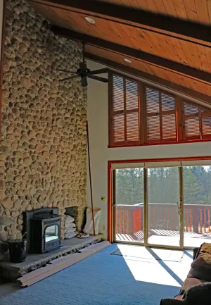 Spacious cabin living room with stone fireplace, wood ceiling, and large sliding glass doors to a snowy deck