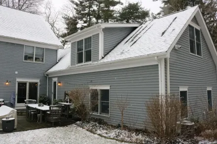 Snow-dusted gray house with a steep roof, patio furniture, and bare shrubs in a winter yard