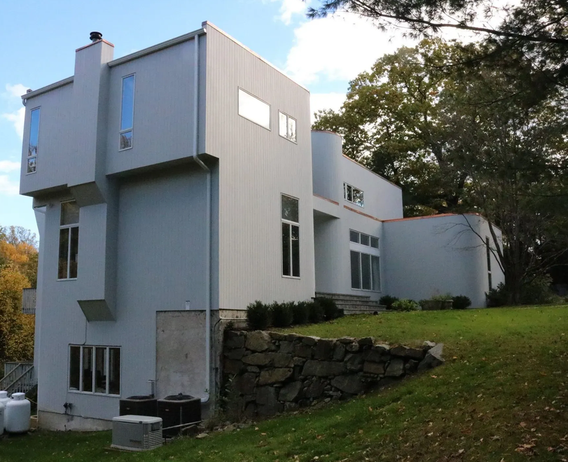 Modern white house with gray trim beside a grassy slope and stone retaining wall, framed by trees