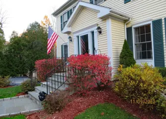 House entrance with American flag, white siding, black shutters, and red bushes along the front steps