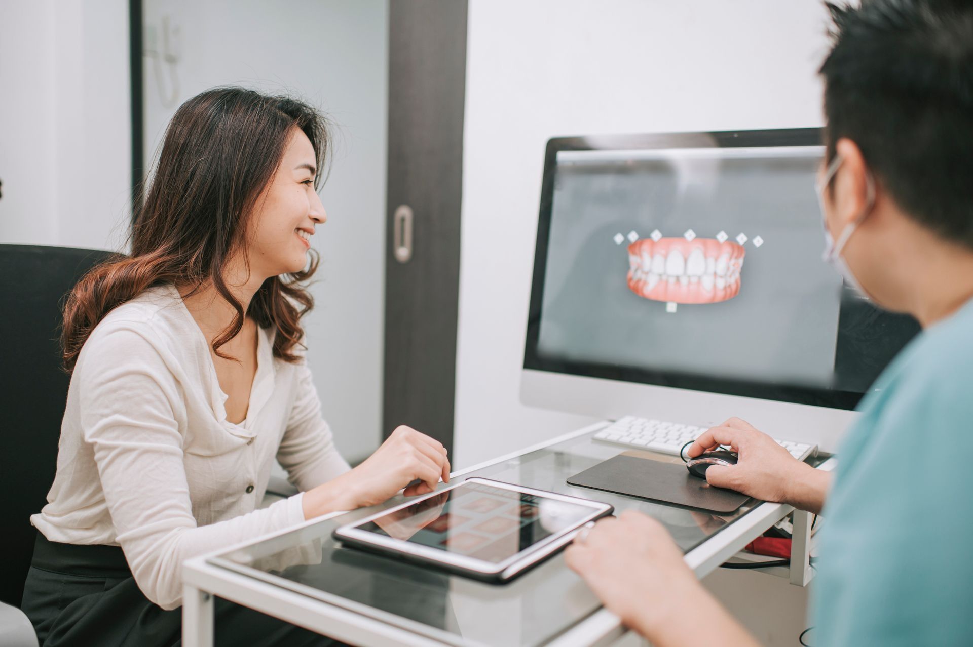 Due persone che parlano sedute a un tavolo di vetro in un ufficio moderno, con un modello dentale su un monitor alle loro spalle.