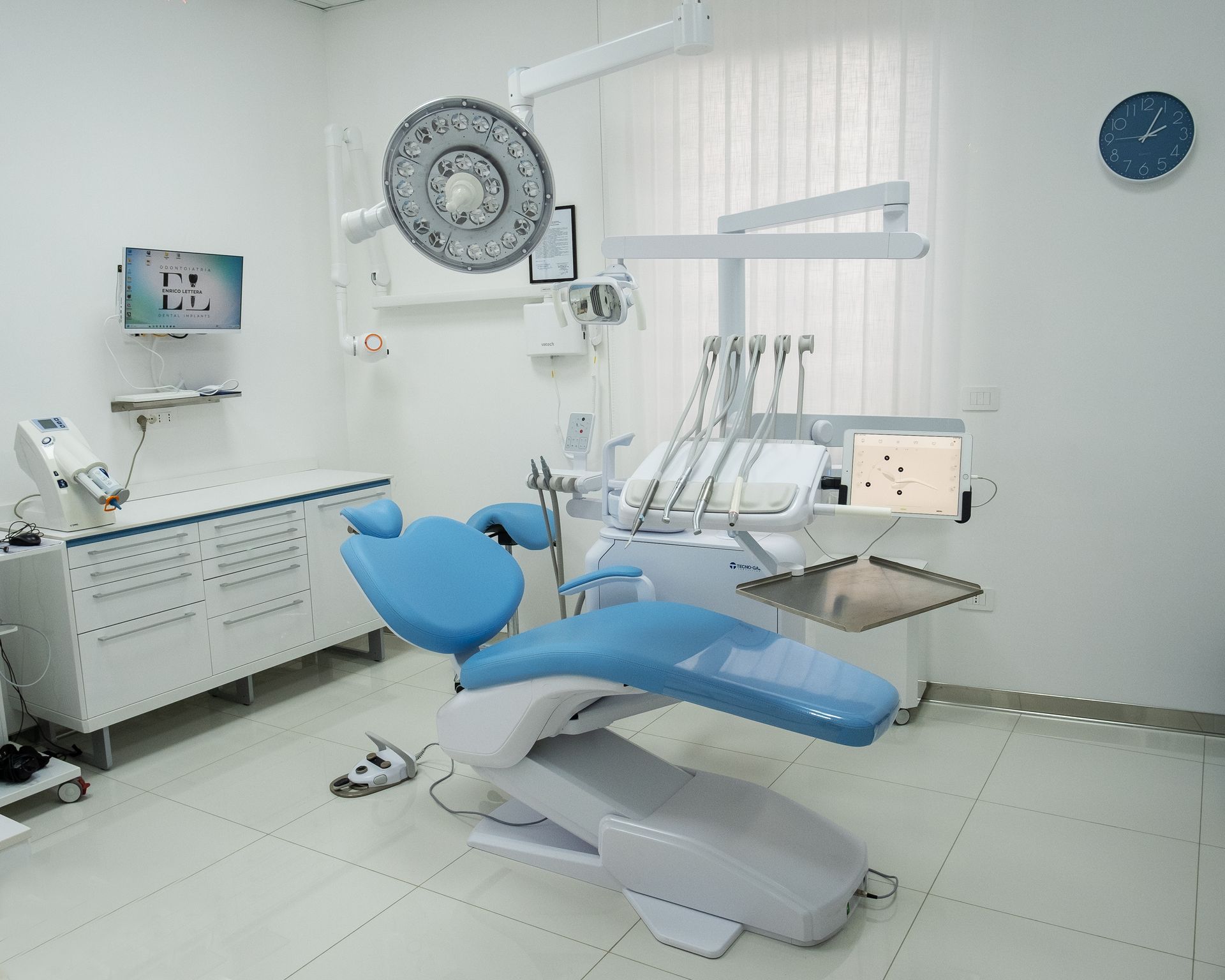 Poltrona odontoiatrica blu in una clinica luminosa e moderna, con strumenti dentali, armadietti e un orologio a parete.