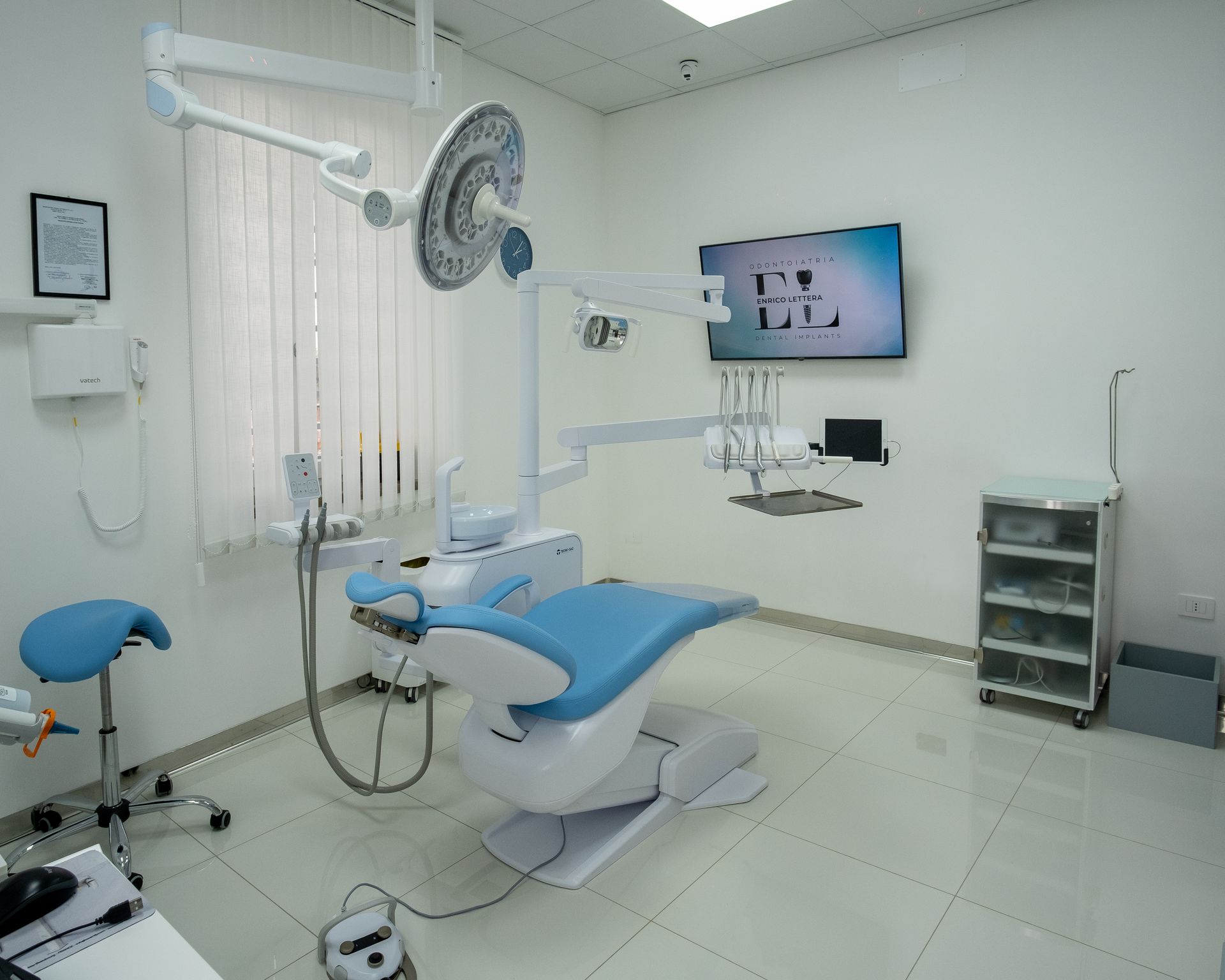Moderna sala per visite odontoiatriche con poltrona blu, strumenti, monitor a parete e armadietti bianchi luminosi.