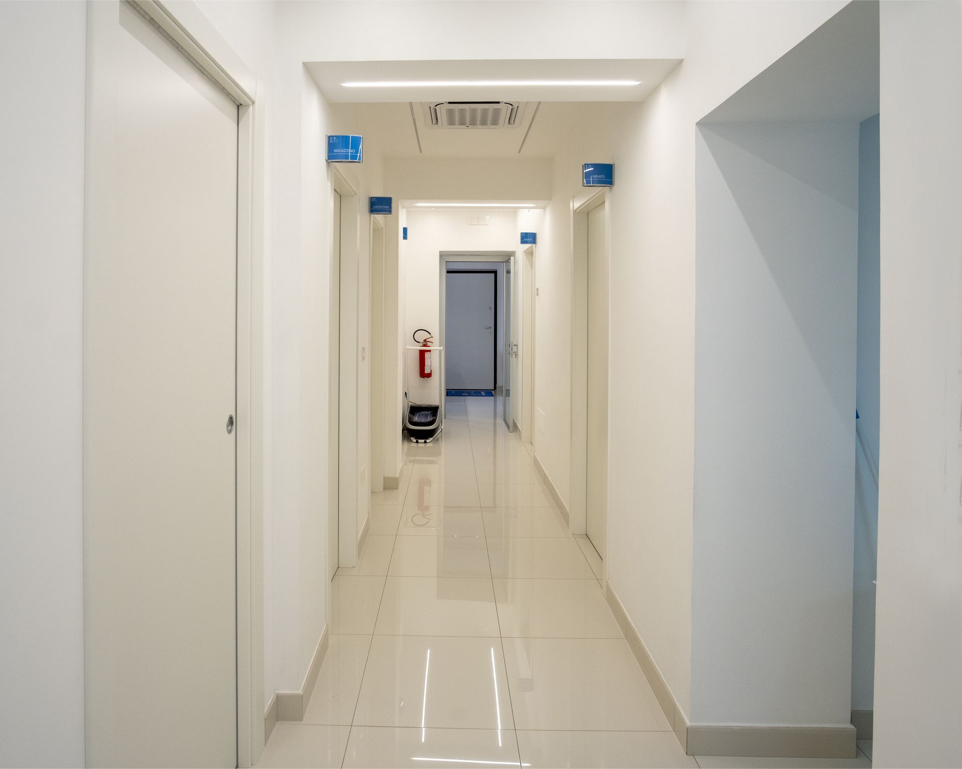Un corridoio bianco e luminoso con pavimento in piastrelle lucide, porte su entrambi i lati e un estintore in lontananza.