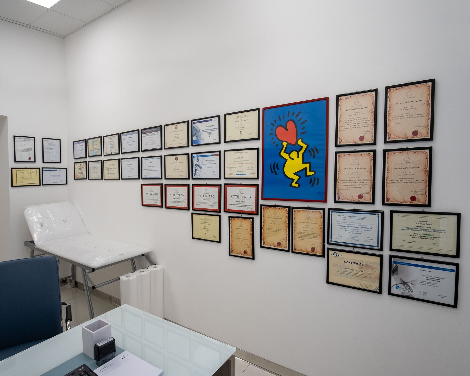 Una parete ricoperta di certificati incorniciati e opere d'arte in un ufficio bianco, con un poster blu al centro.