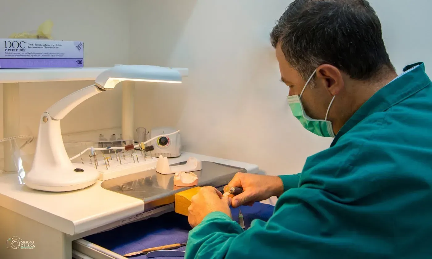 Tecnico in camice verde al lavoro su un banco di laboratorio con un modello dentale sotto una lampada da lavoro