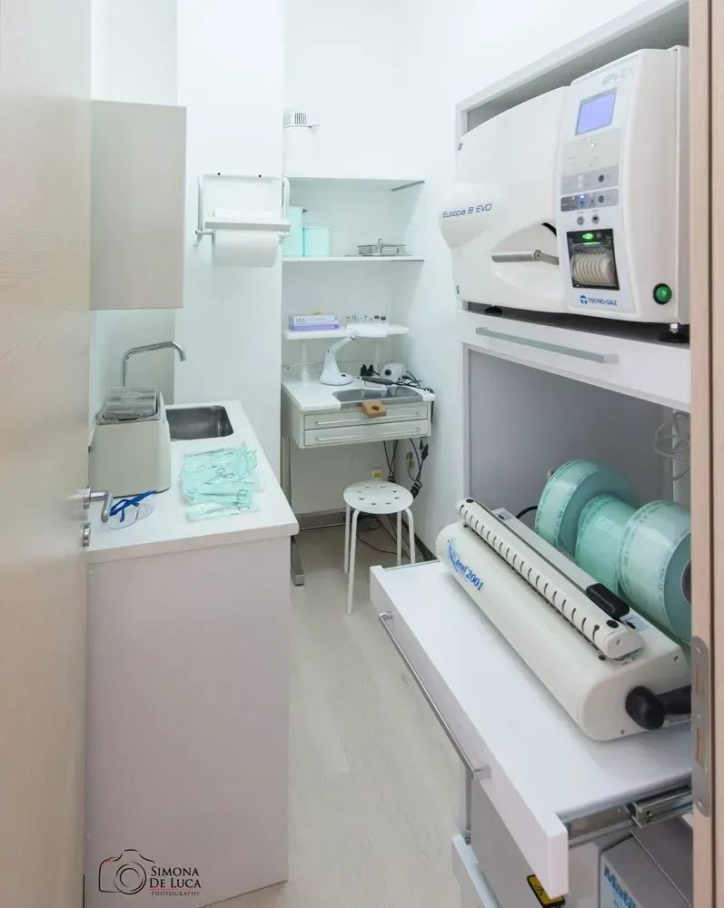 Sala di sterilizzazione dentale con autoclave, lavandino, scaffali e vassoi per strumenti confezionati singolarmente.