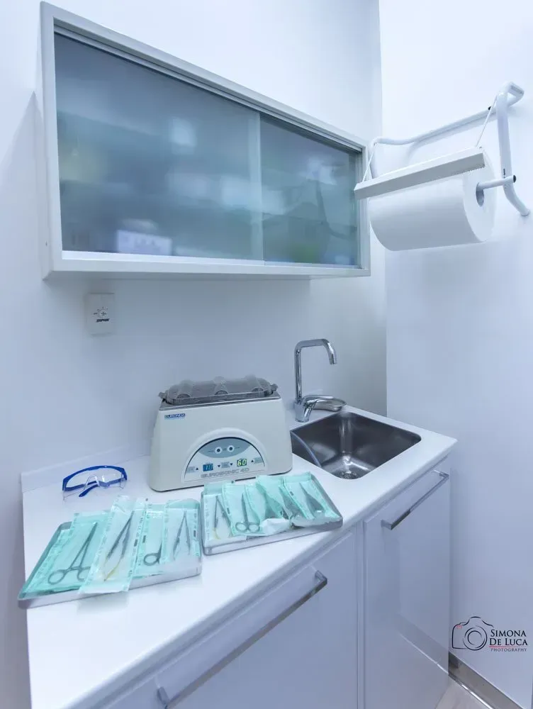 Sala per trattamenti odontoiatrici sterile con lavandino, sterilizzatore, armadietti e strumenti confezionati su un bancone.