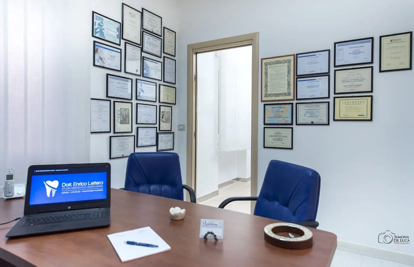 Sala conferenze con certificati incorniciati, sedie blu e un computer portatile su un tavolo di legno.