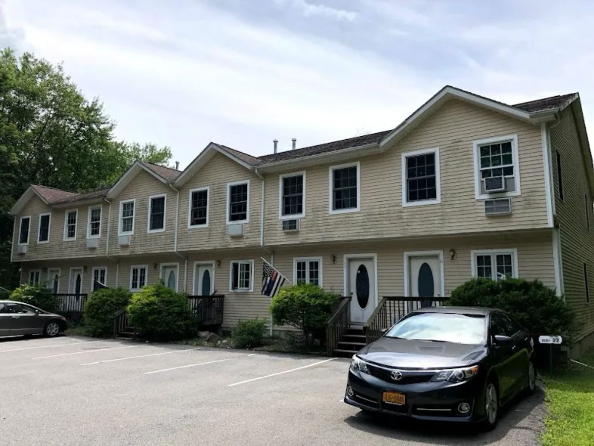 Apartment building with tan siding, dark windows, and parked cars on a paved lot.