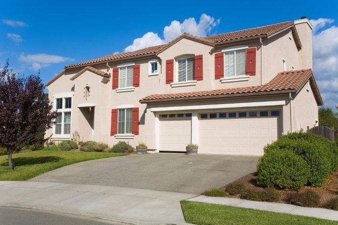 Suburban two-story house with red shutters, white garage doors, and a driveway under a blue sky