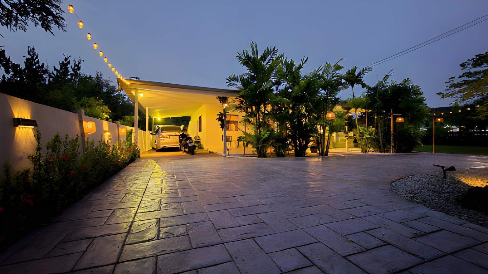 Well-lit driveway and patio at dusk with a parked car, trees, and warm string lights
