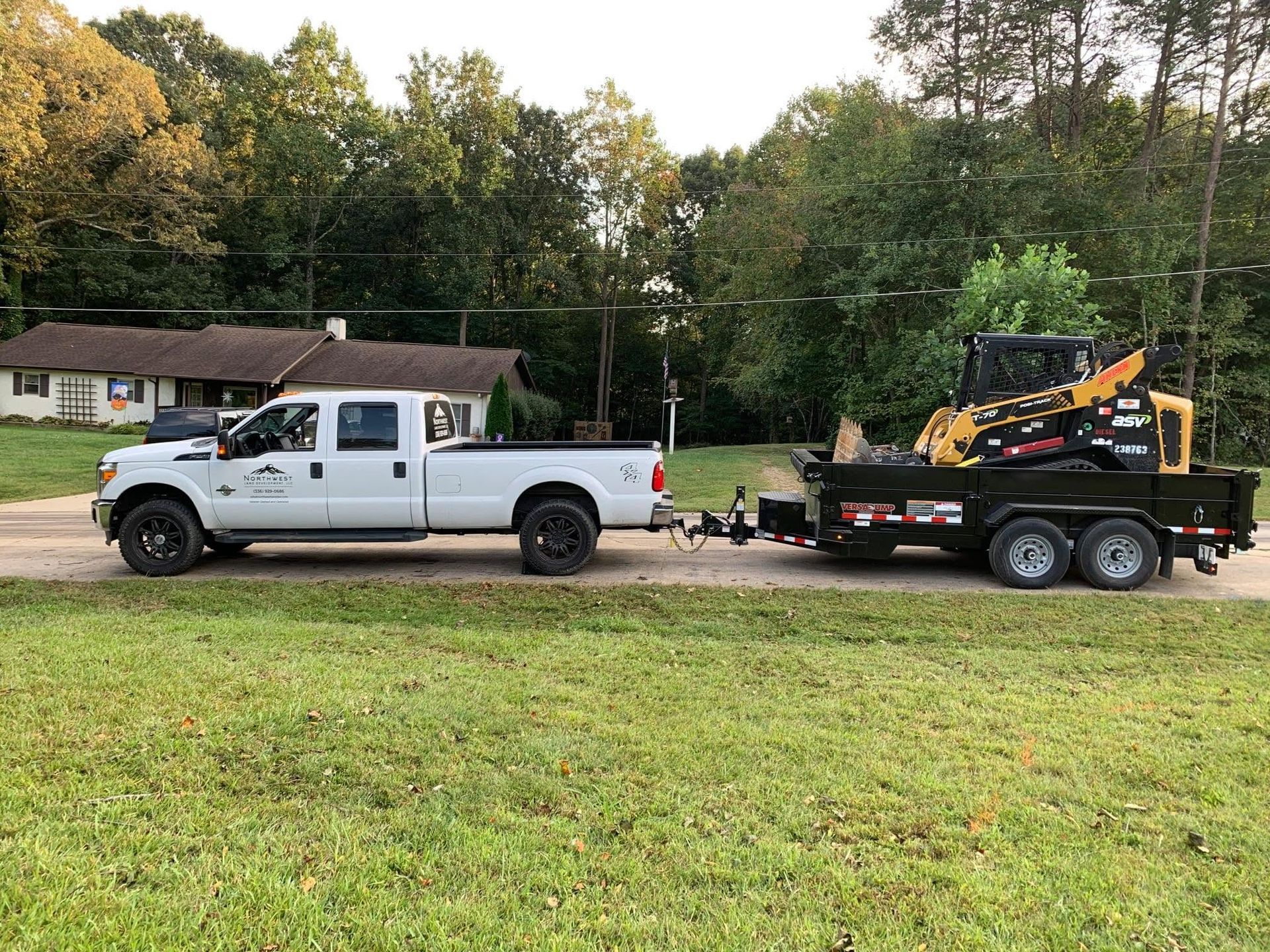 White pickup truck towing a black equipment trailer with a yellow excavator on a roadside near houses and trees