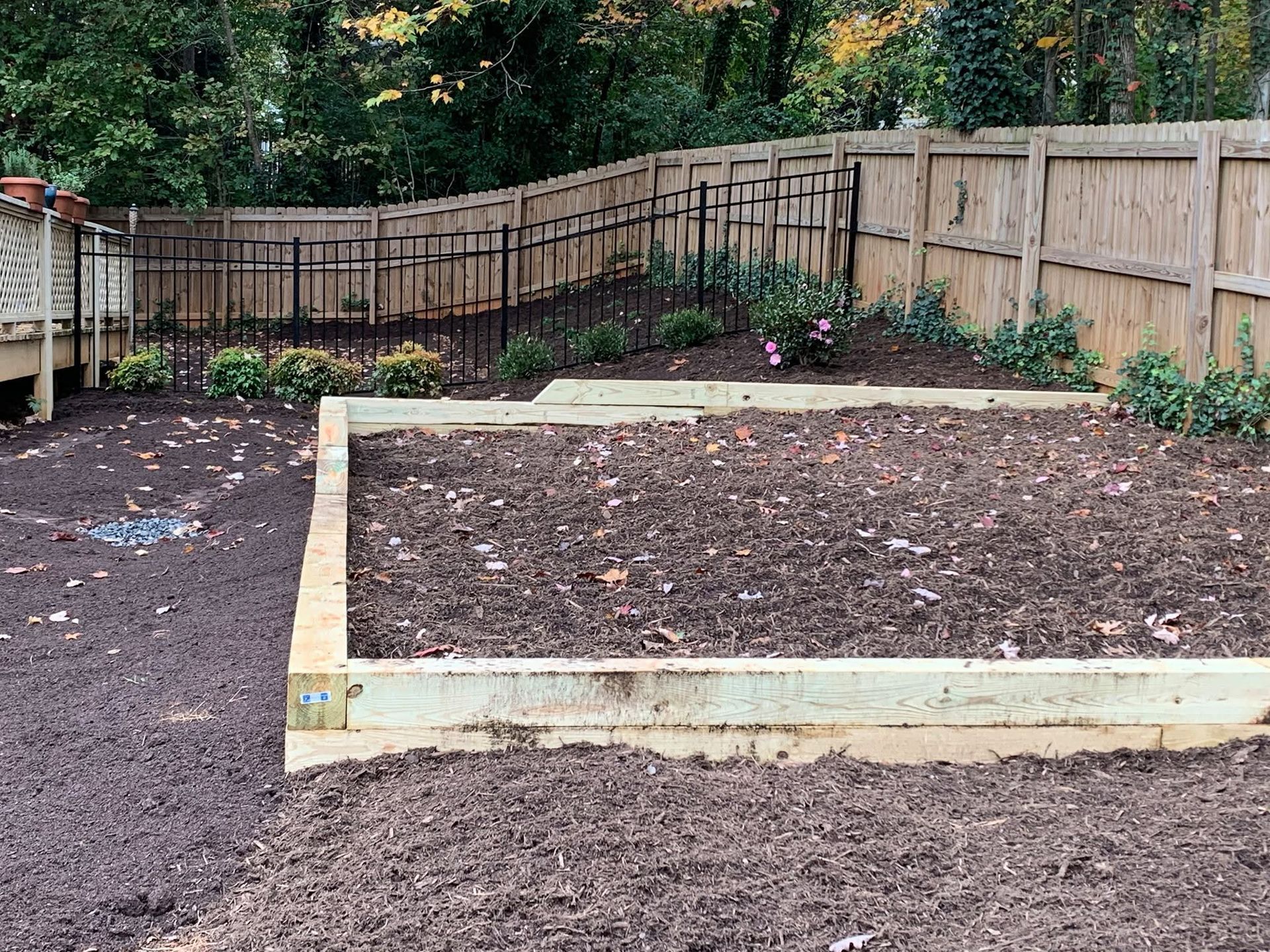 Raised garden bed in a backyard with fresh soil, bordered by a wooden fence and trees.