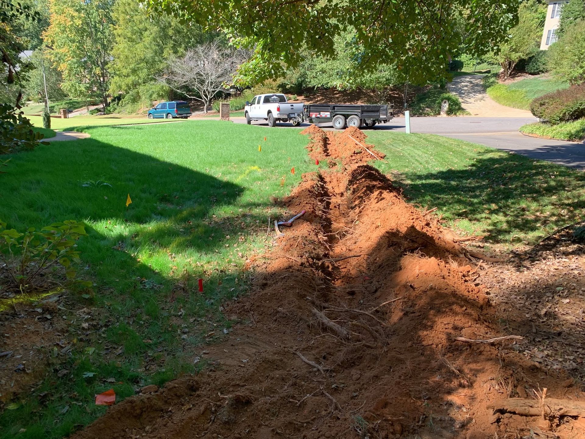Fresh dirt trench cut through a grassy yard beside a road with parked cars and trees.