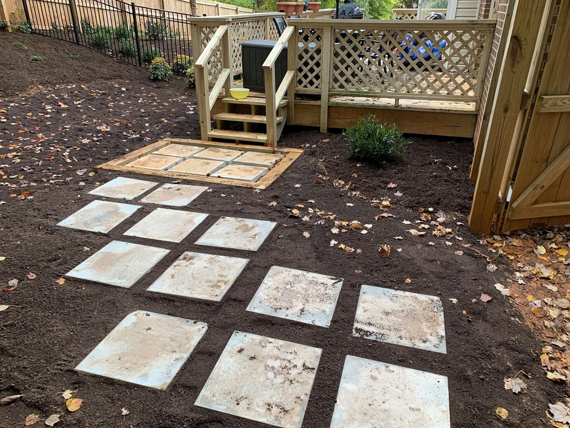Backyard patio with square stone pavers leading to a wooden deck and steps, surrounded by mulch and a fence