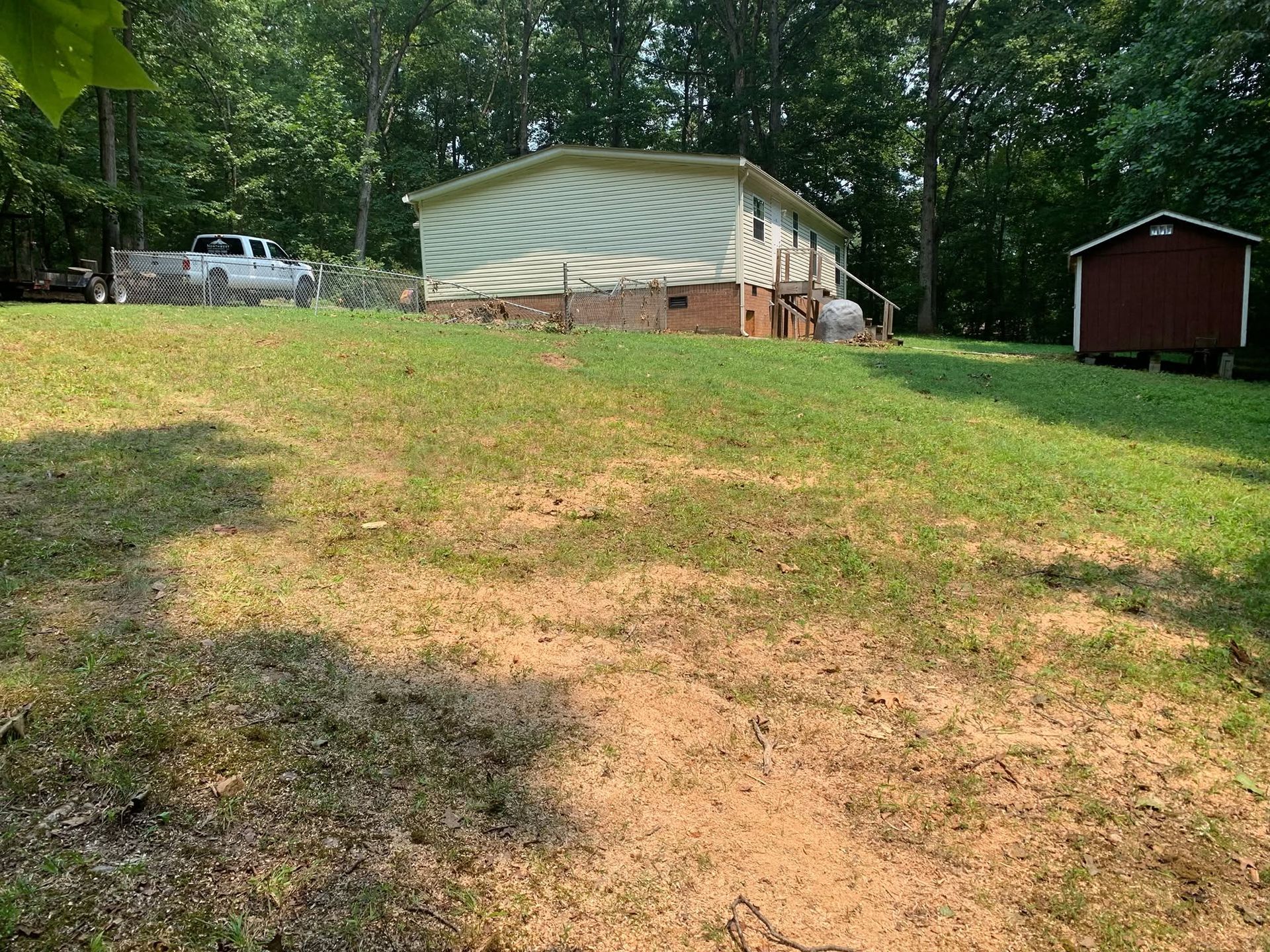 Grassy hillside leading to a beige mobile home, with a small red shed and trees in the background.