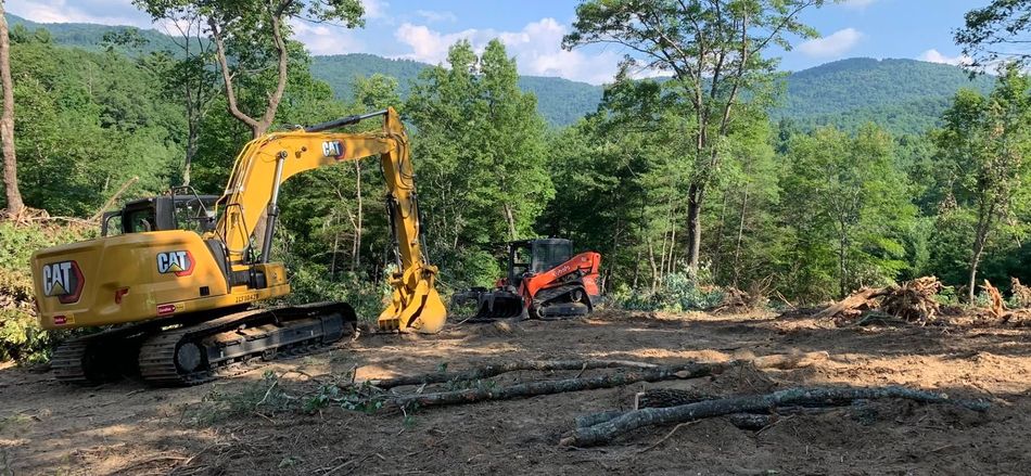 Yellow excavator and orange skid steer clearing a wooded site with mountains in the background