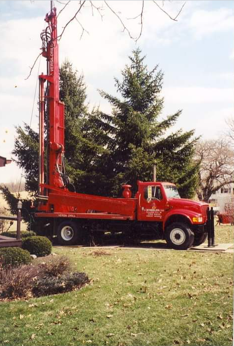 Red drilling rig on a truck, parked in a yard with evergreen trees in the background.