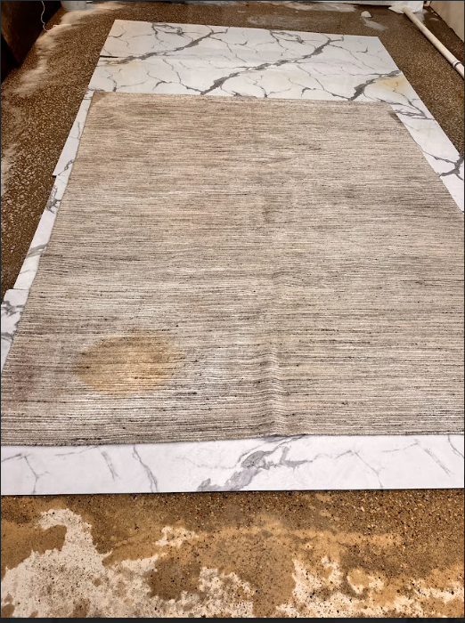 Textured beige rug on a marble-look tile floor with a water stain near the bottom edge.