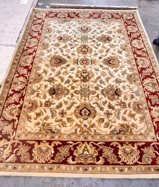 Beige floral patterned rug with red border laid on a concrete floor.