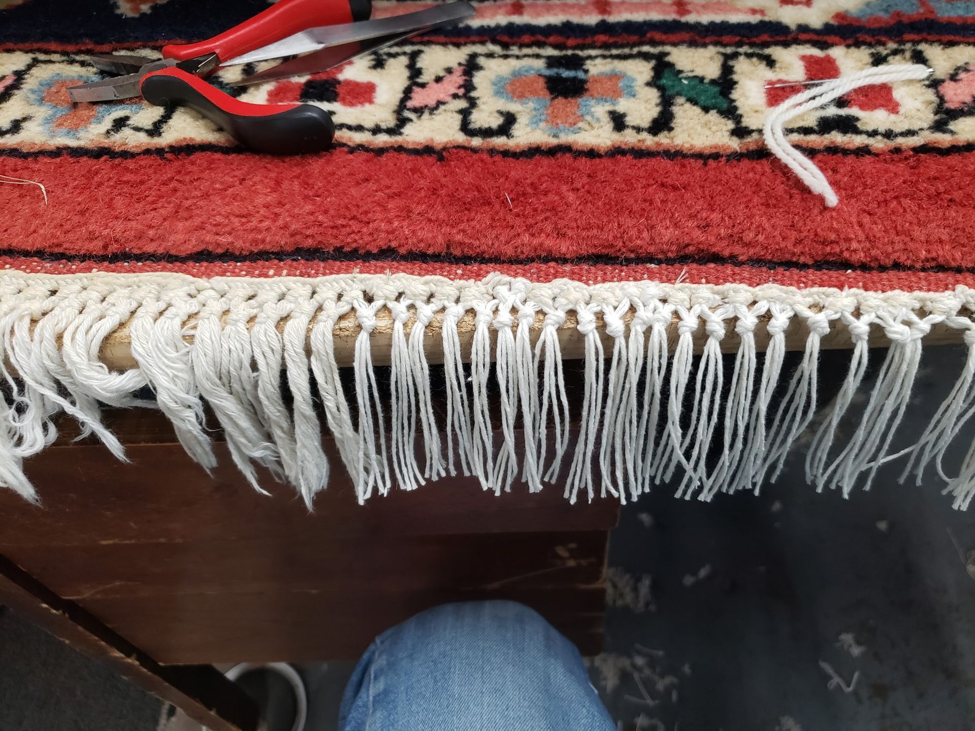Red patterned rug with white fringe on a dark wooden table, with black and red pliers on top.
