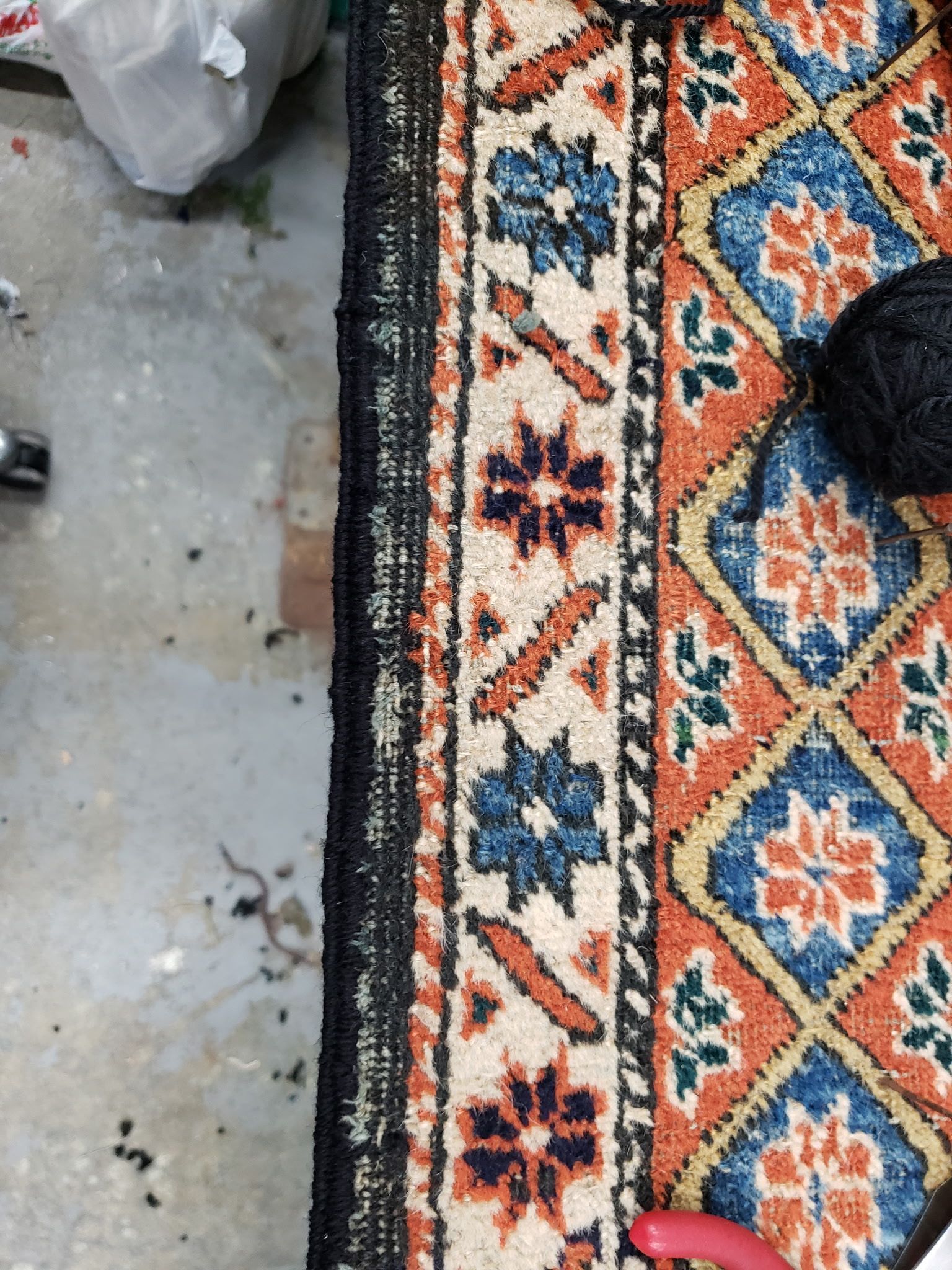 Colorful patterned rug beside a concrete floor, with a black shoe at the edge and a white plastic bag nearby.